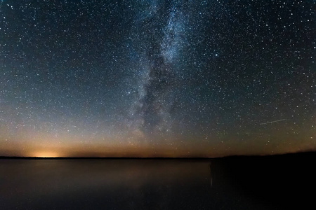 蓝夜星空，银河系在湖泊和俄罗斯村庄上方。 夜晚闪耀的星星