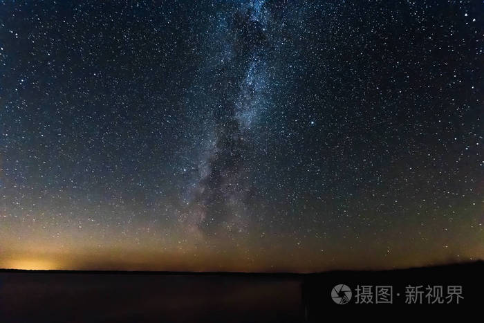 蓝夜星空,银河系在湖泊和俄罗斯村庄上方. 夜晚闪耀的星星