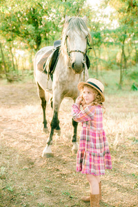 一个漂亮的小女孩，穿着一件老式的格子连衣裙，戴着一顶草帽，还有一匹灰色的马，浅卷发。 秋天的农村生活。 马和人