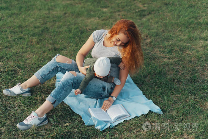 美丽的妈妈正在和她可爱的小男孩读一本书,坐在草地上微笑着