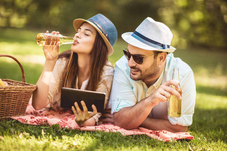 一对夫妇躺在公园里的野餐毯上，野餐篮里装满了水果，他们正在使用数字平板电脑