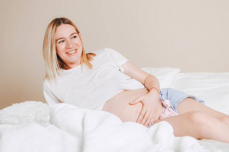 怀孕母亲护理生活方式概念鲍勃发型金发女人保健。 白光室内妈妈坐在床上抱着袜子和肚子。 带壁炉的房间