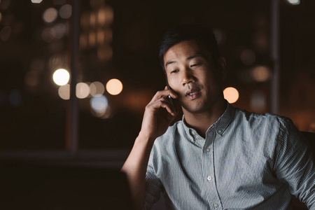 深夜手机年轻的亚洲商人深夜独自一人在办公室工作,在手机上聊天,背景