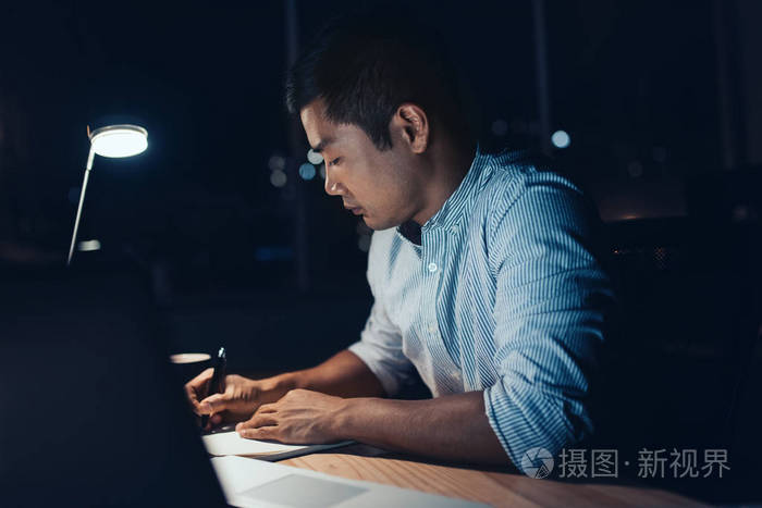 年轻的亚洲商人坐在办公桌前写笔记,晚上在黑暗的办公室里用笔记本