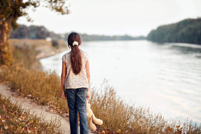 女孩带着玩具熊在日落时沿着森林中的小路走着美丽的河流和风景