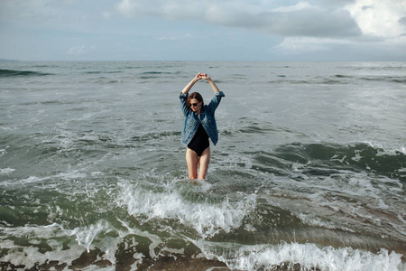 快乐微笑黑发女人穿着黑色泳装和牛仔夹克在海洋背景下享受在海洋中行走，举起双手。巴厘岛，仓古，印度尼西亚
