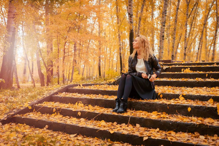 那个女孩坐在秋天森林的楼梯上。 黑发女人穿着皮夹克，手里拿着手机短信和上网。 黄叶落下。
