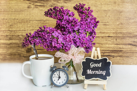 早安健康带有闹钟,咖啡杯和木制背景上的早安短信的丁香花束.