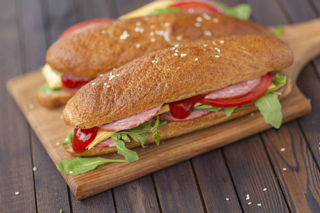 两个面包三明治与萨拉米奶酪，阿鲁古拉，沙拉，西红柿和洋葱在一个切割板。 深色木制背景上的三明治。 美味的小吃。