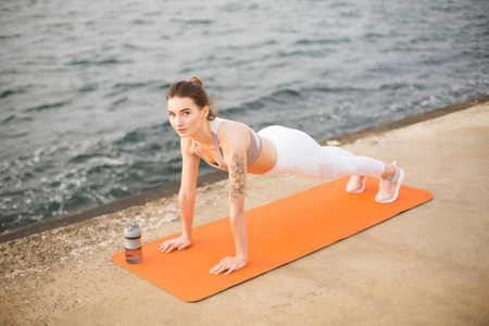 年轻的女人梦想地看着相机在海边练习瑜伽。 漂亮的女孩，穿着运动上衣和白色紧身裤，在橙色瑜伽垫上做运动锻炼，背景海景