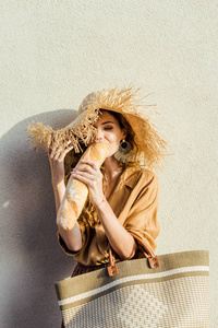美丽的年轻女子戴着草帽，站在白墙前，吃着面包