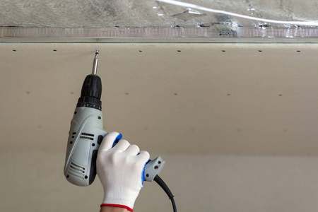 关闭施工工人手中的保护手套与无电缆螺丝刀连接悬挂的干壁天花板到金属框架。 改造建设和DIY理念。