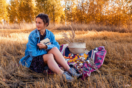 秋季生活方式和家庭户外娱乐的概念。 在新鲜空气中野餐一个年轻的女人穿着牛仔夹克和连衣裙，用野餐篮苹果酒把书放在格子上。 在背景