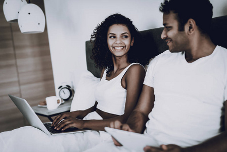 美国黑人夫妇看着床上的笔记本电脑。 早晨休闲。 休息一下。 看屏幕上。 在沙发上不寒而栗。 家里的衣服。 假期休息。 一起微笑。