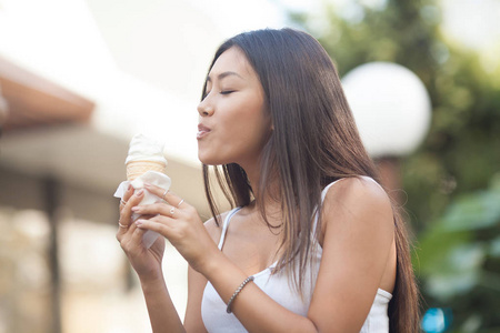 在公园的绿树背景下，快乐的年轻微笑的亚洲黑发女人在吃冰淇淋。