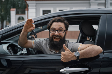 汽车经销商是最幸福的客户。 有魅力的男人拿着车钥匙，竖起大拇指，牙齿微笑，坐在车里，透过窗户看镜头。