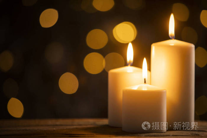 用模糊的圣诞灯光背景,在木桌上合上三支燃烧的蜡烛照片-正版商用图片