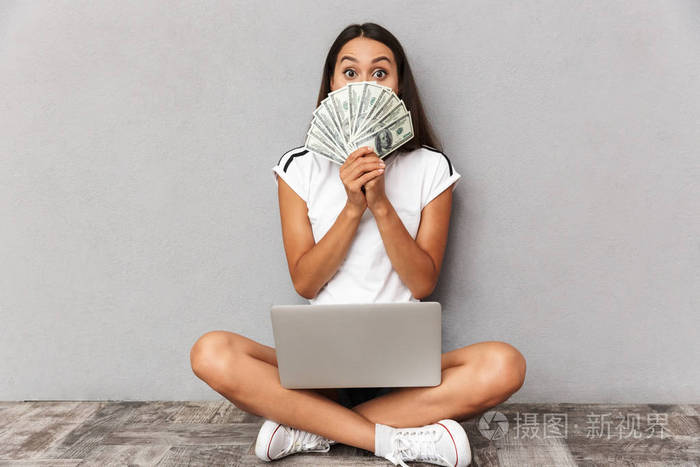 兴奋的图像震惊的年轻女子坐在灰色背景上使用笔记本电脑拿着钱遮住脸