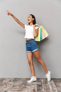照片的年轻快乐的女人拿着购物袋孤立在灰色背景指向。
