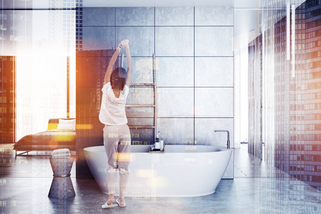 穿着睡衣的女人站在豪华浴室里，灰色瓷砖墙混凝土地板白色浴缸和靠近墙壁的木梯。 主卧室在后台。 色调图像双曝光