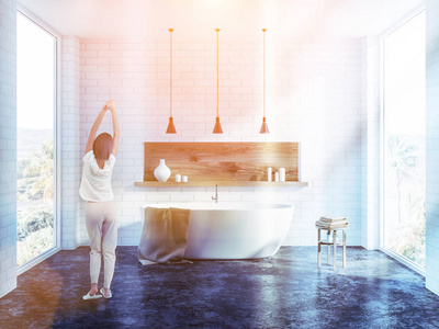 早上在白色砖浴室内部伸展的女人，里面有一个混凝土地板，一个白色浴缸，几个天花板灯和一个木制架子和蜡烛。 调色图像