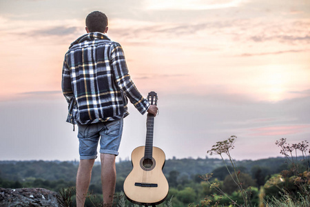 那家伙背着吉他站在大自然的背景上,夕阳美丽照片