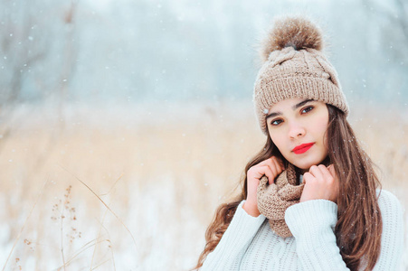 冬天特写美丽微笑的年轻女人的肖像，穿着针织帽子和毛衣，在烟雾下在户外行走。 涂红唇膏的女孩很暖