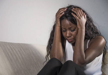 年轻有吸引力和悲伤的黑人非裔美国妇女坐在沮丧的家里沙发上，感到焦虑和沮丧，患有抑郁问题焦虑危机或周期性疼痛