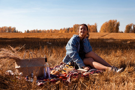 秋季生活方式和家庭户外娱乐的概念。 一个年轻的女人穿着牛仔夹克，穿着在格子花格子上享受大自然的衣服，拿着野餐篮苹果酒。 在背景秋