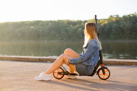 在日落时，金发女人穿着短裙和牛仔裤坐在滑板车上。 放松旅行度假概念