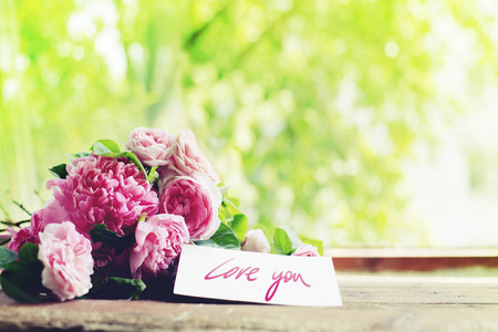 室外木制桌子上放着一束带爱你卡片的粉红色牡丹玫瑰花束