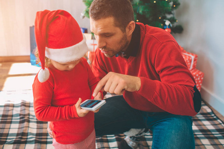 圣诞快乐, 新年愉快。父亲和女儿的图片一起。孩子手持电话。人展示和技术的点。孩子戴着圣诞帽。他们在树与箱子