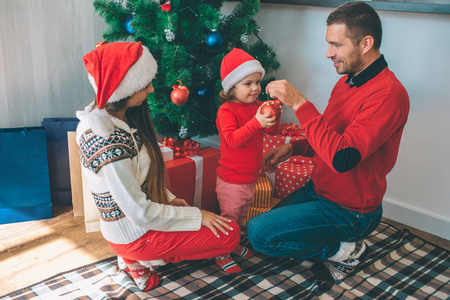 圣诞快乐, 新年愉快。幸福的家庭坐在一起在圣诞树上。年轻人拿着红色玩具, 小女孩有整件事。她全神贯注。女人看他们