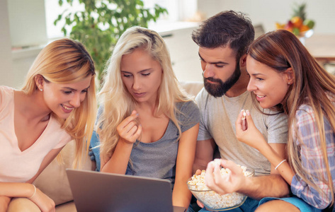 一群开朗的朋友看着笔记本电脑，在家吃爆米花。