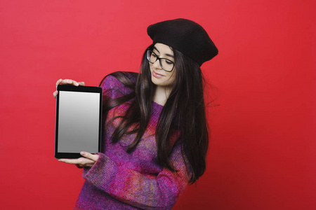 漂亮的年轻女士穿着时尚的服装和眼镜，微笑着展示了带空白屏幕的现代平板电脑，同时站在明亮的红色背景下
