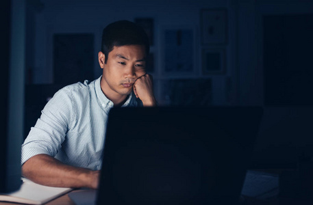 傍晚时分，一个疲惫的年轻亚洲商人坐在办公桌前用笔记本电脑在黑暗的办公室里工作