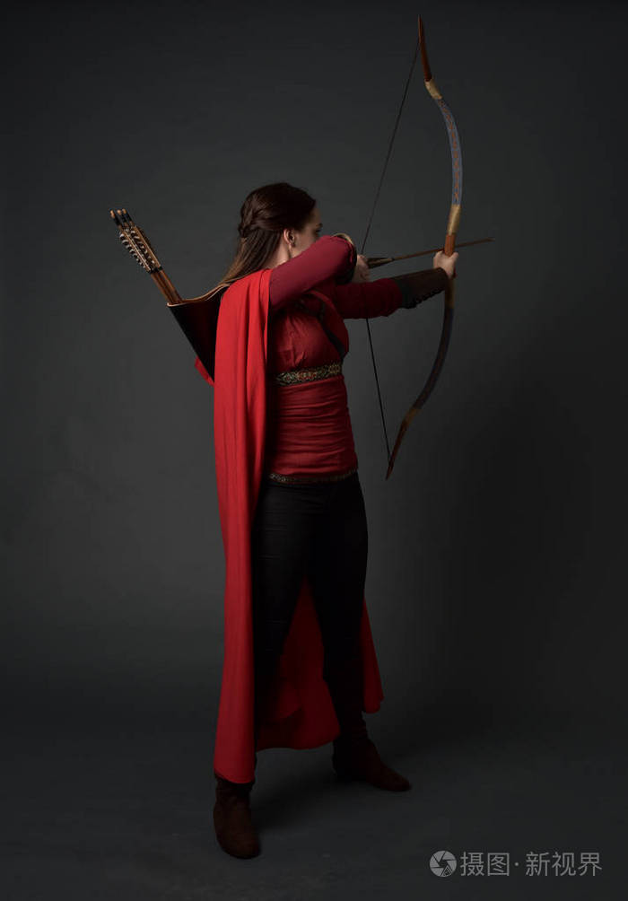 穿着红色中世纪服装和斗篷的黑发女孩的全长肖像手持弓箭站在灰色工作