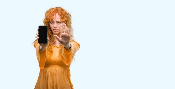 年轻的红发女人，用张开的手展示智能手机，做停止标志，严肃自信的表情防御手势