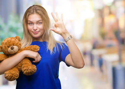 年轻的白种人女人抱着可爱的泰迪熊在孤立的背景下做OK标志与手指优秀的符号