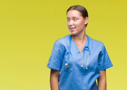 年轻的白种人医生女人穿着医疗制服，在孤立的背景上看着一边，脸上带着微笑，自然的表情。 自信地笑着。