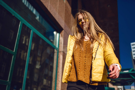 一位年轻的欧洲妇女带着长长的金发，戴着黄色夹克，黄色太阳镜，走在市中心街道上拍摄。 即使是光。
