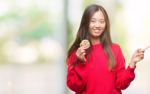 年轻的亚洲女人在孤立的背景上吃巧克力片饼干，非常高兴地用手和手指指向一边