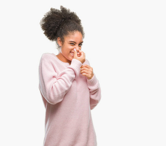 年轻的非洲美国女人穿着冬天的毛衣，在孤立的背景上闻到一些臭味和令人恶心的难以忍受的气味，用手指在鼻子上屏住呼吸。 难闻的概念。