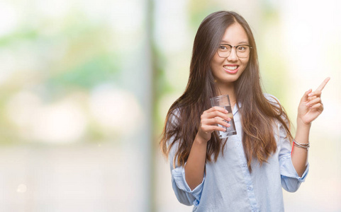 年轻的亚洲女人喝着一杯水，在孤立的背景下，非常高兴地用手和手指指向一边