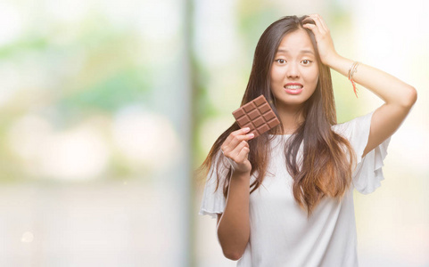 年轻的亚洲女人在孤立的背景下吃巧克力棒，用手压在头上，羞愧和惊讶的脸，愤怒和沮丧。 因为错误而恐惧和不安。