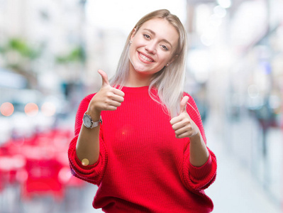 年轻的金发女人穿着冬天的毛衣，在孤立的背景上，批准用手竖起大拇指做积极的手势，微笑着，为成功而高兴。 看着相机赢家的手势。