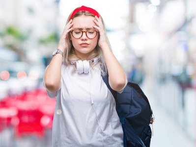 年轻的金发学生妇女戴着眼镜和背包，在孤立的背景下，因为疼痛和偏头痛而感到头痛绝望和压力。 手放在头上。