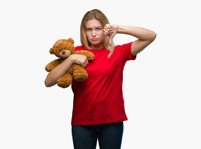 年轻的白种人女人抱着可爱的泰迪熊，在孤立的背景下，愤怒的脸，消极的迹象，表现出厌恶，拇指向下拒绝的概念