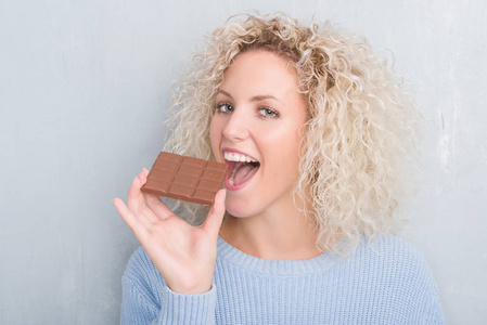 年轻的金发女人在灰色的墙上吃巧克力棒，聪明的脸上有自信的表情，严肃的思考
