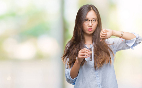 年轻的亚洲女人喝了一杯水，在孤立的背景上，愤怒的脸，负面的迹象，显示出厌恶，拇指向下拒绝的概念